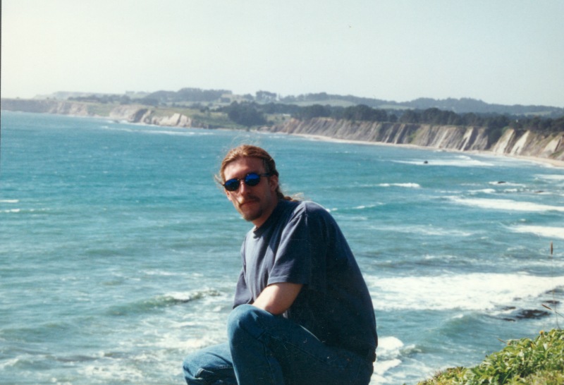 Brian W Root crouching with Nothern California coastline behind him.